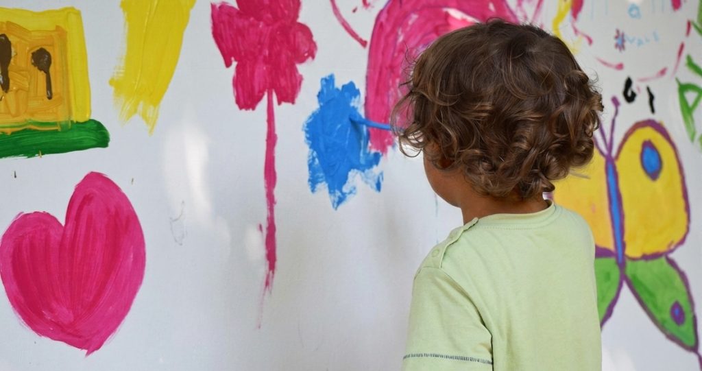 Ein Kind malt bunte Motive auf die Wand, in einer Kita.