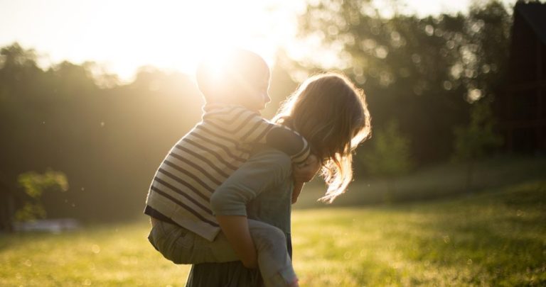 Großes Mädchen trägt ihren kleinen Bruder auf dem Rücken. Sie stehen auf einer Wiese